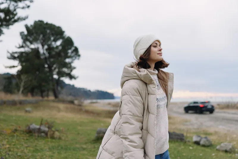 Woman in a warm coat stands outdoors on a breezy field near the shore, creating a serene landscape with soft light and distant traffic.