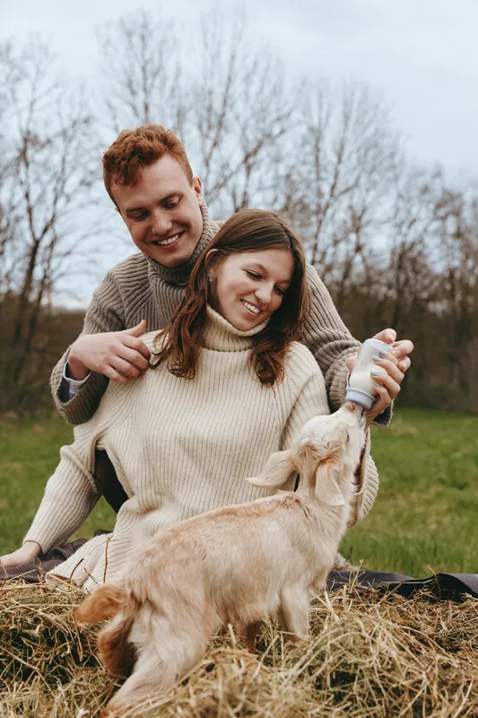 Smiling couple feeding a friendly goat kid in a field, cozy sweaters, outdoor moment showcasing lifestyle and companionship, soft natural light, rustic setting, warm mood, candid care, play