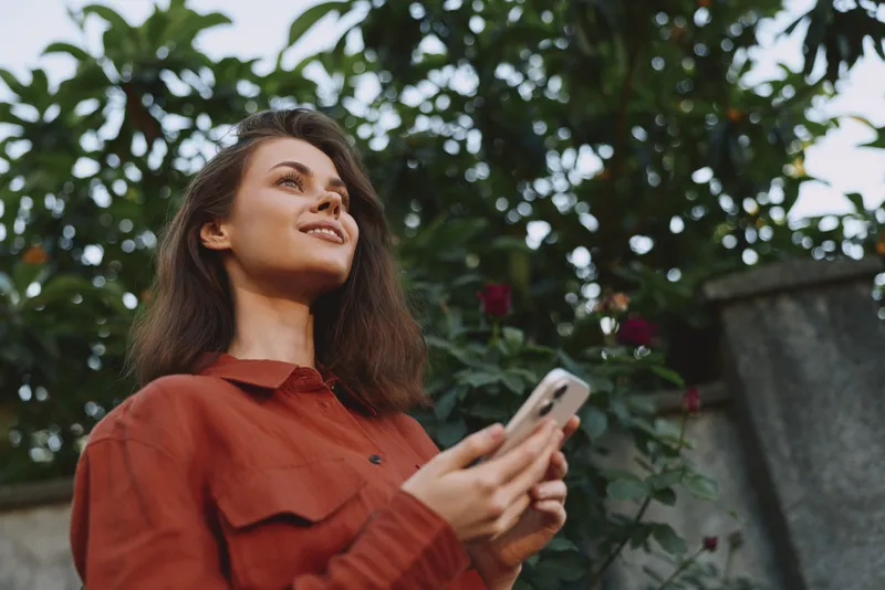 Young brunette woman in a red shirt stands outdoors, looking up with a smile while holding a phone, surrounded by lush garden greenery and soft daylight, creating a casual lifestyle moment
