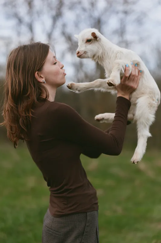 Young woman with long brown hair holds a small white baby goat close to her face outdoors in a natural setting, showing affection and connection on a cloudy day.
