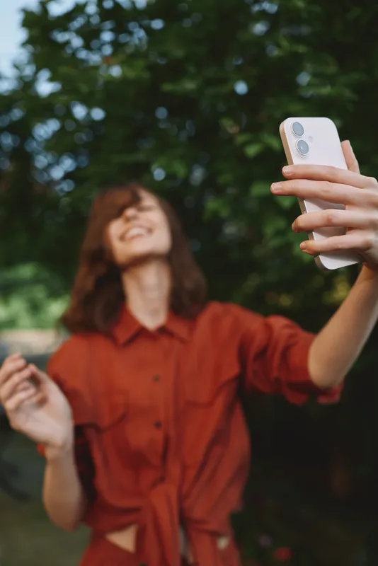 Smiling woman selfie outdoors smartphone lifestyle happiness casual portrait nature wearing red shirt as she snaps a moment with a phone. Warm daylight creates a friendly, candid mood