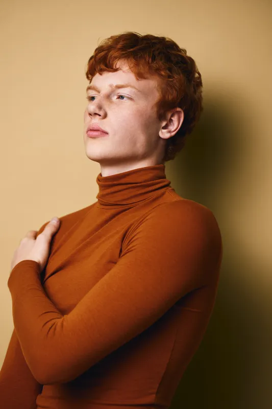 Portrait of young man with red hair wearing brown turtleneck sweater looking away. Studio shot with plain background showing thoughtful expression and casual style clothing.