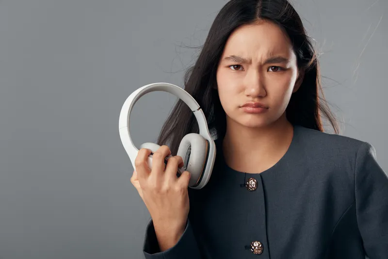 Young woman with long dark hair holding wireless headphones with displeased expression on gray background. Frustrated female listening device audio technology accessorry.