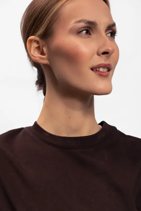 Portrait of a young woman with clear skin and natural makeup wearing a dark brown sweater looking to the side. Studio shot on white background with soft lighting and neutral expression.