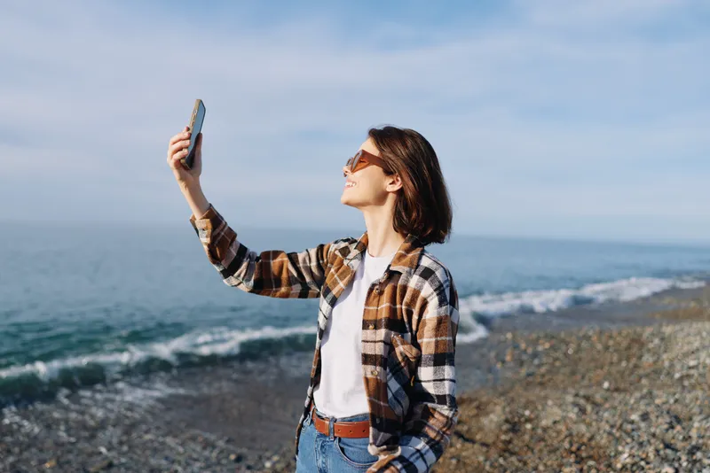 Woman on a beach wearing a plaid shirt and sunglasses takes a selfie with a phone by the ocean, casual lifestyle shot against the blue sky and rocky shoreline.