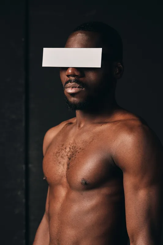 Creative conceptual portrait of shirtless African man with paper strip covering eyes in dark studio. Visual style emphasizes skin texture and artistic anonymity in modern photography.