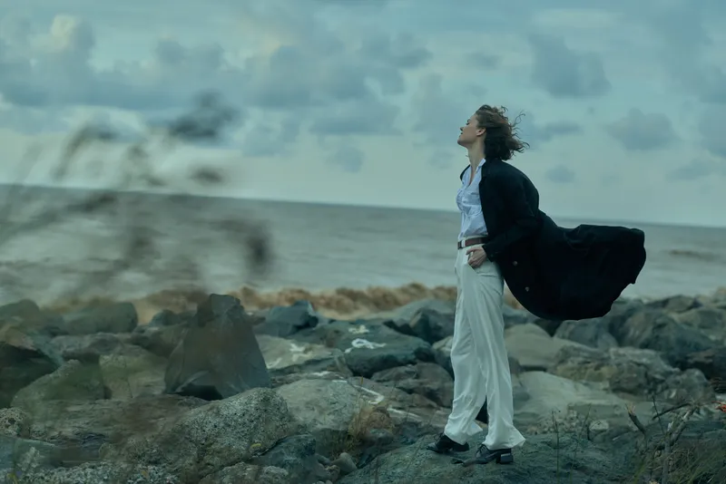 Wind sweeps along the sea and coastline as a woman strides on rugged rocks in a flowing coat and white trousers, horizon visible, movement and drama in a bold outdoor fashion moment by the ocean