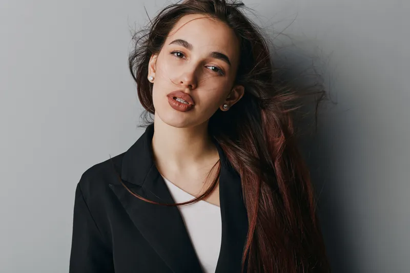 Young woman with long brown hair wearing black jacket and white top standing against gray background looking at camera with natural expression and loose hair strands.