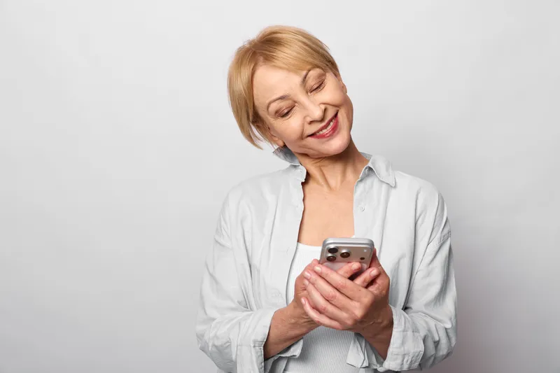 Senior woman holding phone, eyes closed, soft smile and laughter, light studio background, relaxed posture, communication with mobile technology, calm copy space, today