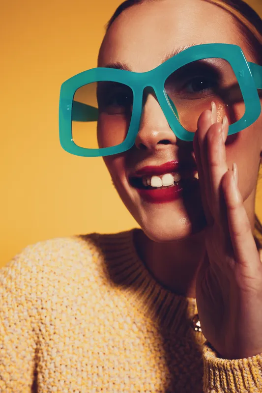 Close-up of smiling woman wearing large turquoise glasses and yellow sweater on yellow background. Concept of fashion, style, joy, and colorful accessories in studio lighting.