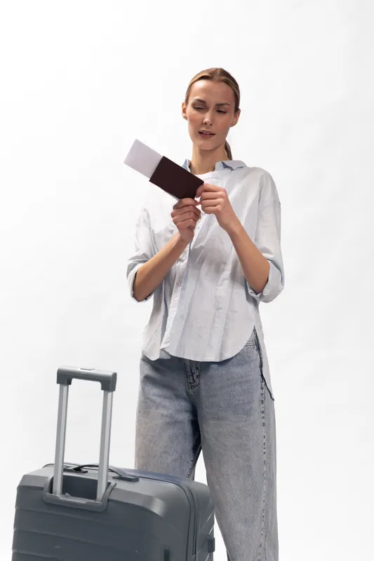 Young woman in casual clothes holding passport and tickets stands near suitcase on white background. Concept of travel, vacation planning, and airport preparation.