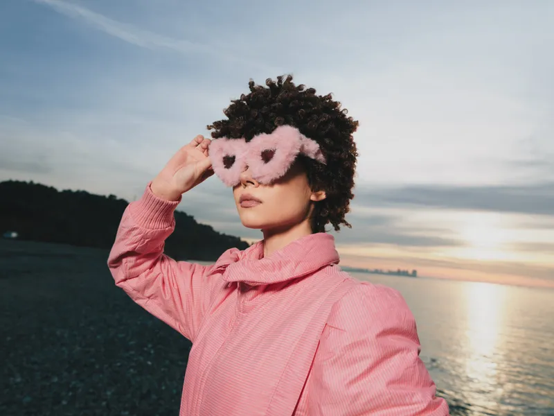 Woman wearing a pink coat and heart shaped sunglasses stands at a sunset beach, creating a bold fashion moment with dreamy outdoor mood, soft light and calm water in the background