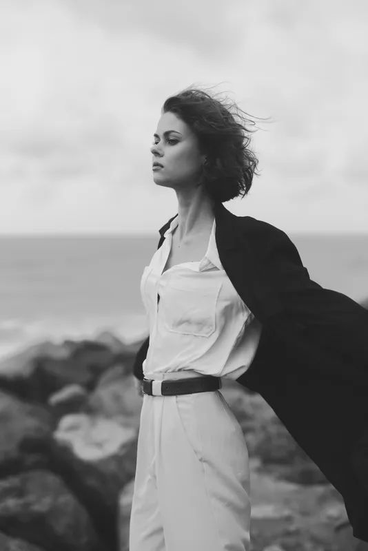 Woman on a windy beach wearing a white dress and dark coat, standing with arms outstretched, sea in the background, monochrome fashion mood.