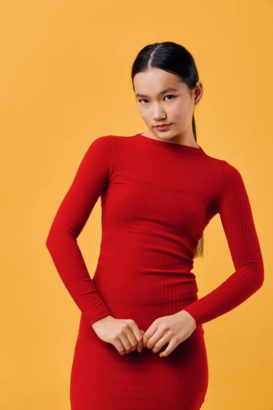 Young woman in red long sleeve dress standing against orange background with confident posture and neat hairstyle. Female model posing with hands clasped in front and calm expression.