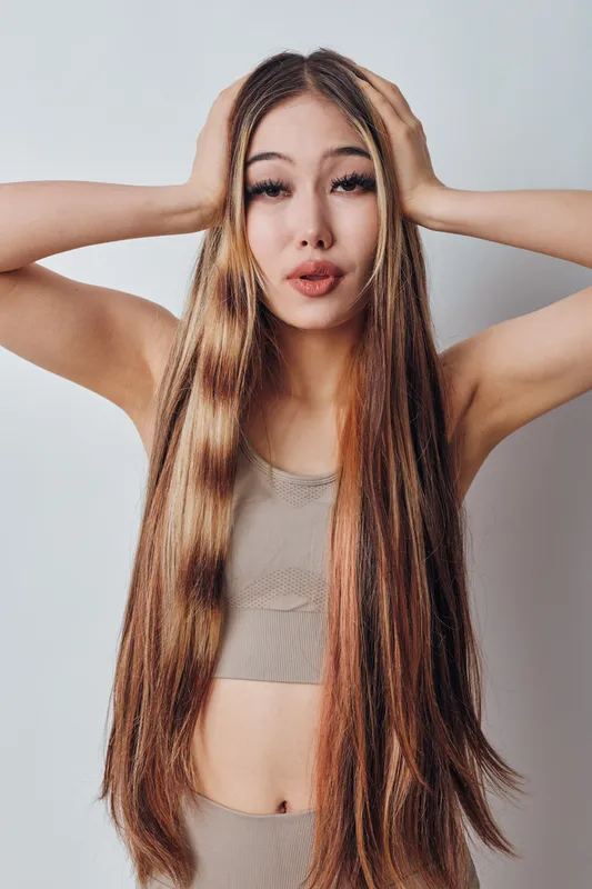 Woman with long straight hair and natural makeup wearing beige crop top holding her head with both hands on white background. Young adult female posing with neutral expression.