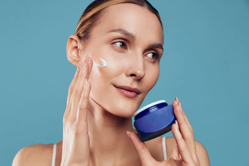 Young woman with blonde hair applies moisturizing cream on her cheek while holding a blue jar against a plain blue background, expressing calmness and self-care.