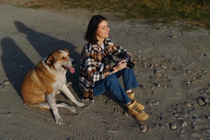 Woman with a camera sits on a gravel path beside a friendly dog, enjoying an outdoor moment. Casual attire, sunlight, and calm scenery celebrate simple companionship.
