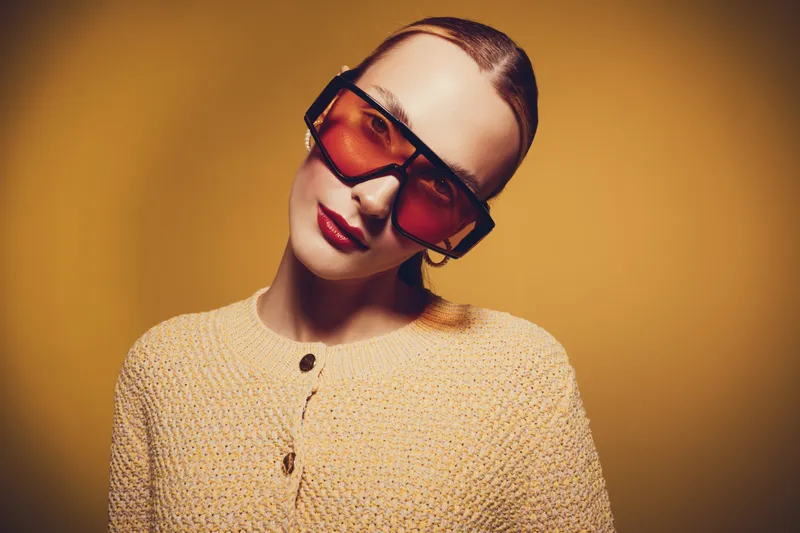 Young woman with slicked back hair wearing oversized red sunglasses and a beige knitted sweater poses against a warm yellow background. Fashion and style concept with confident expression.