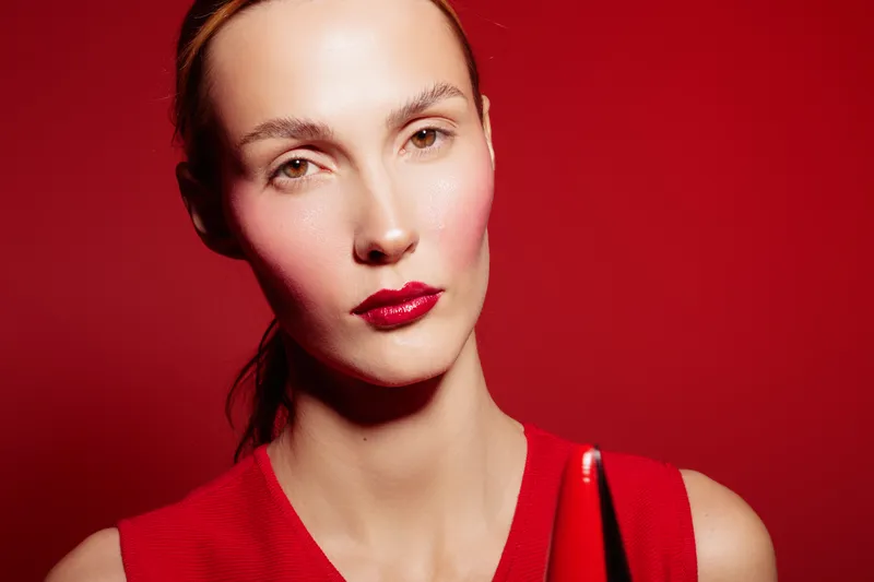 Portrait of a woman with red lipstick and red dress on red background. Studio lighting highlights her clear skin and serious expression. Concept of beauty, fashion, and makeup.