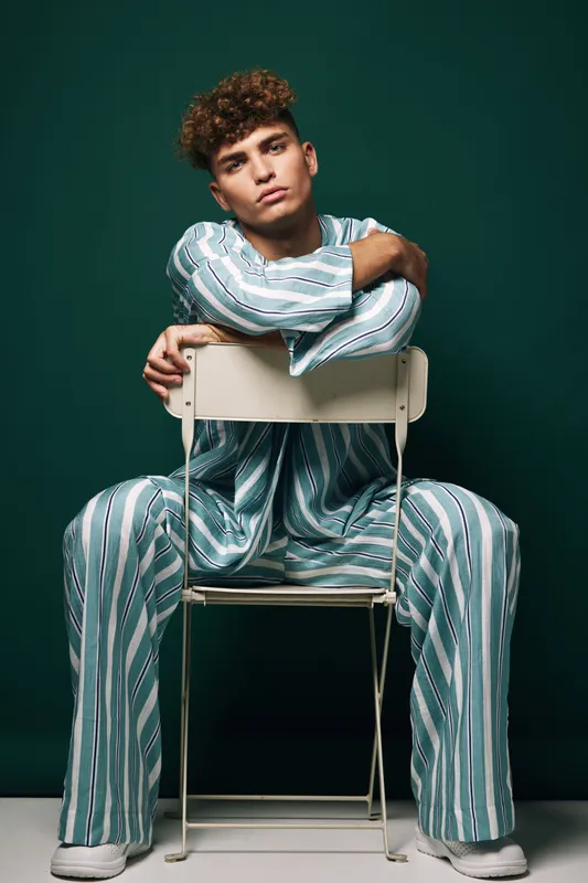 Young man with curly hair wearing striped pajamas sitting on chair backwards against dark green background in studio. Casual fashion, relaxed pose and confident expression.