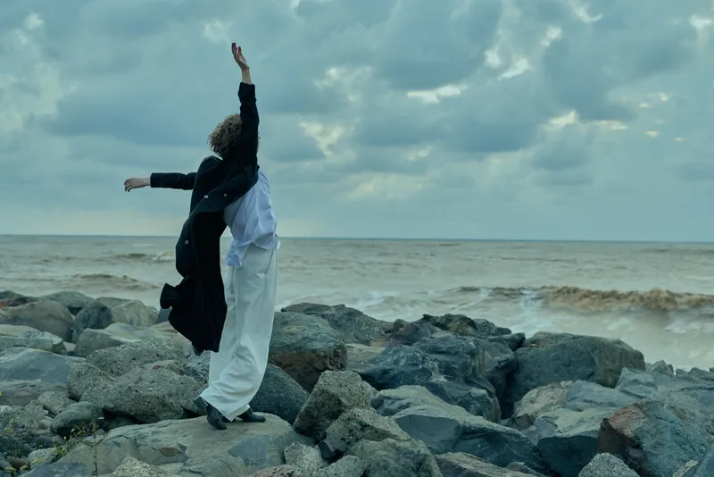 Woman on a rocky shore raises arms toward the open sea, capturing movement, freedom and drama against a moody, windy sky and rugged coast.