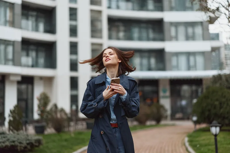 Young woman in a dark trench coat holds a smartphone outdoors, hair blown by the wind, standing in a modern urban courtyard for a candid moment.