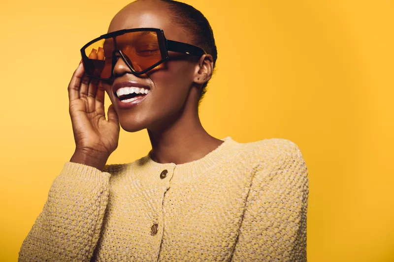 Smiling dark skinned African woman wearing large sunglasses and knitted sweater poses against yellow background. Concept of fashion, style, beauty, and confident female model.