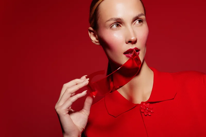 Stylish woman in red coat holds red sunglasses near lips against red background. Fashion portrait with bold makeup and elegant pose. Concept for style, beauty, and color contrast.