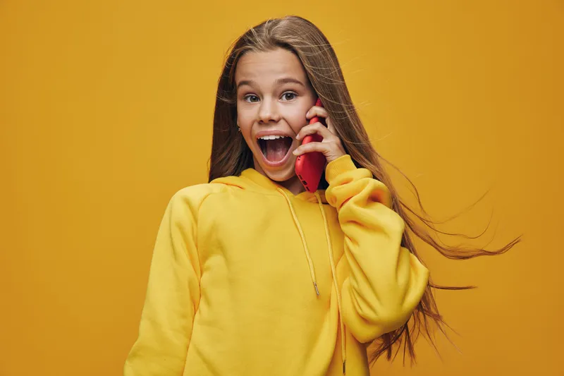 Girl in yellow hoodie talking on red phone with surprised expression on orange background. Child with long hair and casual clothes communicating and excited by call.