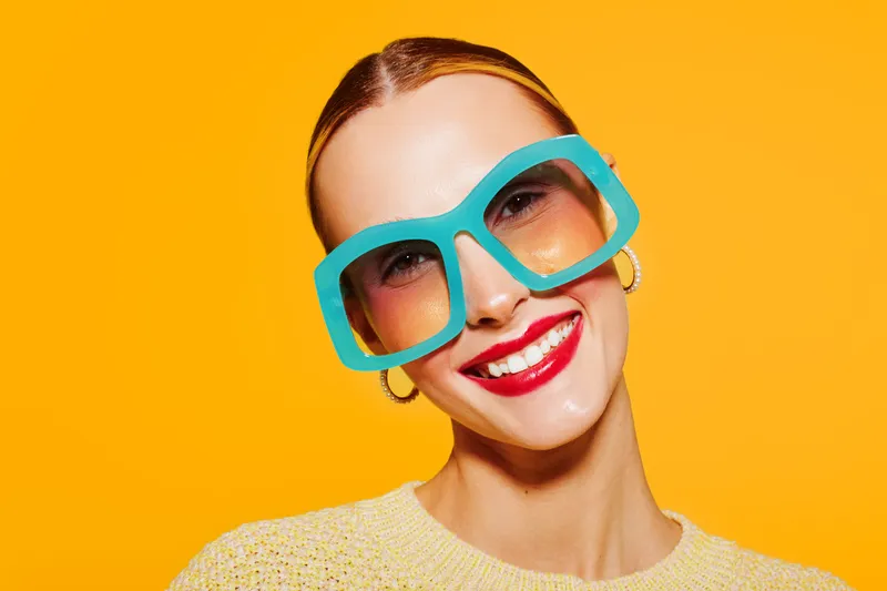 Smiling woman with large blue sunglasses and red lipstick poses against bright yellow background. Fashion portrait with vibrant colors and joyful mood in studio lighting.