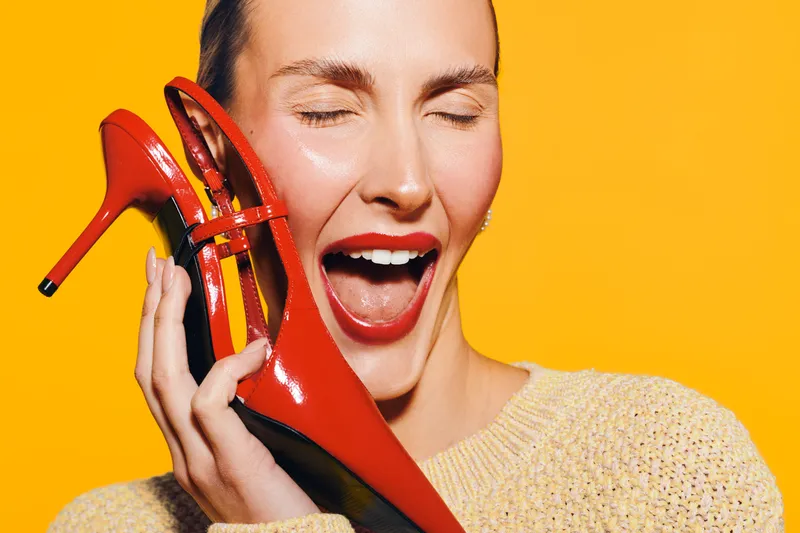 Happy woman with red lipstick holds red high heel shoe near face with closed eyes on bright yellow background. Concept of fashion, style, beauty, and joyful emotions.