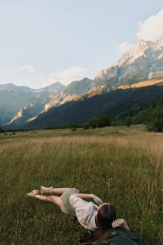 A woman lies in a grassy meadow with distant mountains, enjoying a calm outdoor moment. A lifestyle scene of relaxation and connection with nature.