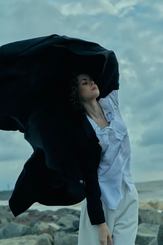 Windy coast scene with a businesswoman lifting a dark coat as a white shirt and blazer billow against a turbulent sky, an outdoor fashion moment conveying motion, confidence, and bold professional