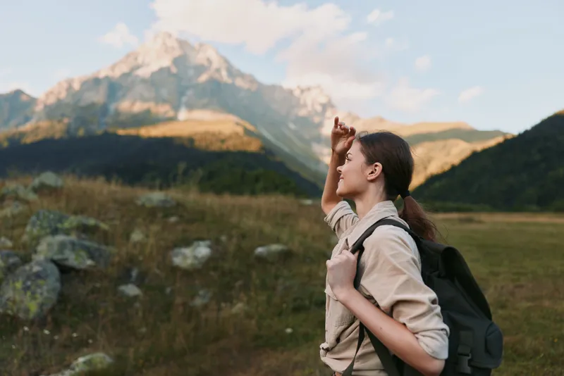 Young woman hiking outdoors in mountain landscape, carrying backpack, looking at distant peak, casual outfit, natural light, adventure and lifestyle concept