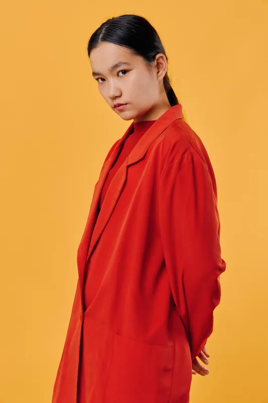 Young woman wearing red coat stands against yellow background with hands behind back looking contemplative and confident in studio portrait fashion concept with minimal style.