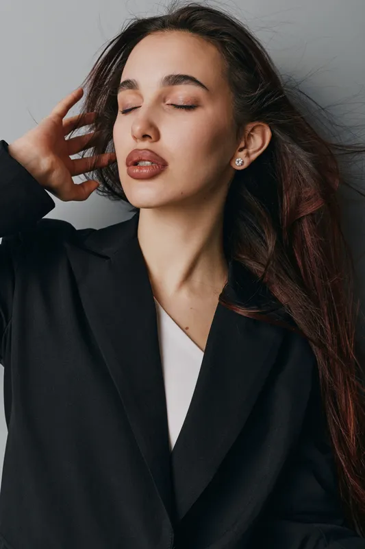 Young woman with long hair wearing black coat posing with eyes closed and hand near head, showing calm expression and natural makeup in studio setting with gray background.