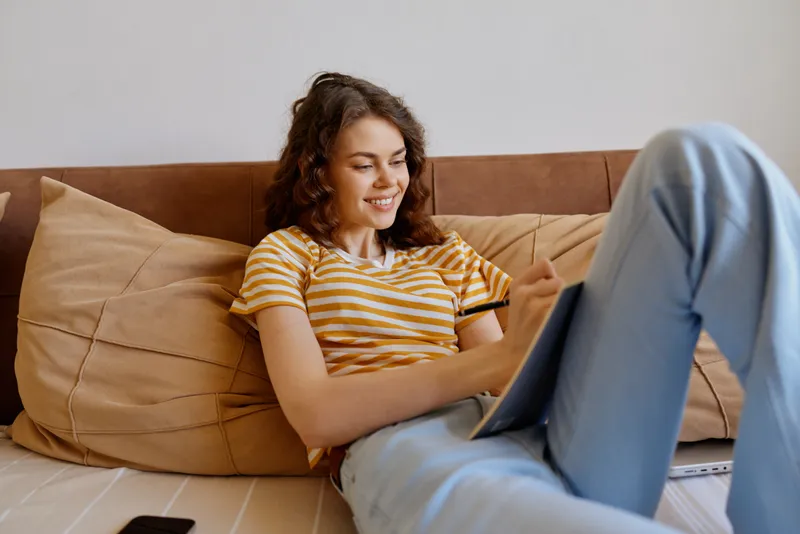 Casual woman lifestyle home, she lounges on a sofa using a laptop, smiling softly, striped tee and jeans, warm natural light in a cozy living room, everyday comfort and focus.