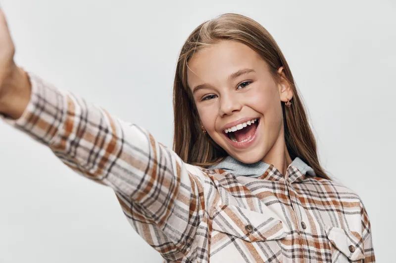 Girl smiling with open mouth and raised arm in studio for happiness, joy, and cheerful expression. Portrait of young female wearing plaid shirt on light background.