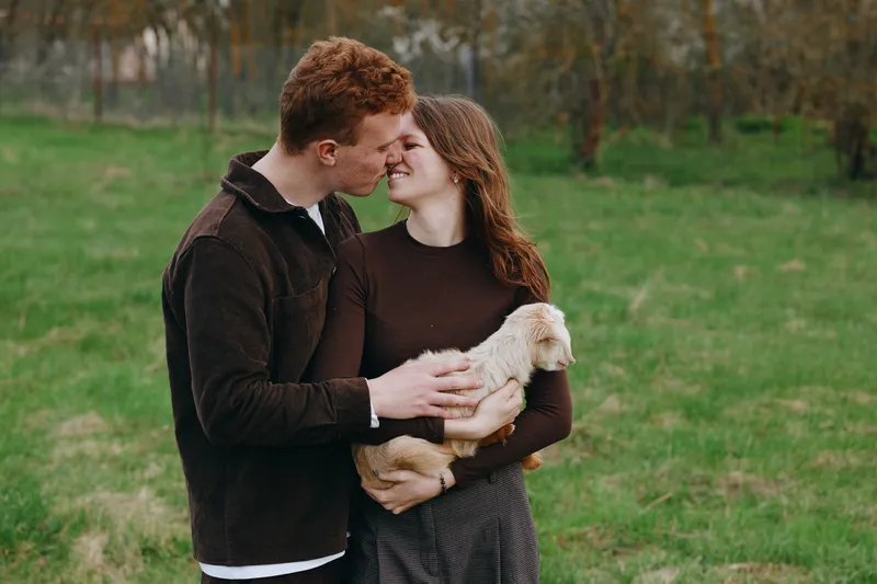 Happy couple in a countryside field cuddle a young goat kid, sharing tender smiles and hands on the small animal, capturing warm lifestyle moment with gentle farm charm.