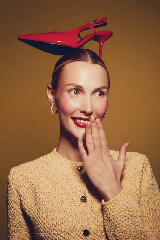Smiling woman with red high heel shoe balanced on her head wears yellow textured sweater and gold hoop earrings. Concept for fashion, style, and playful elegance in studio lighting.