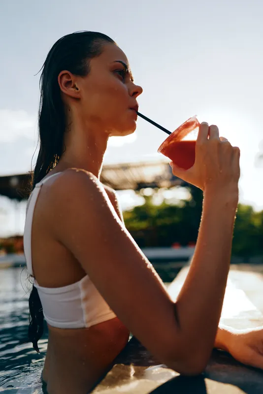 A woman in a white swimsuit sips a drink through a straw by the pool, sunlit water and a relaxed mood. A lifestyle moment of leisure, refreshment, and outdoor summer downtime.