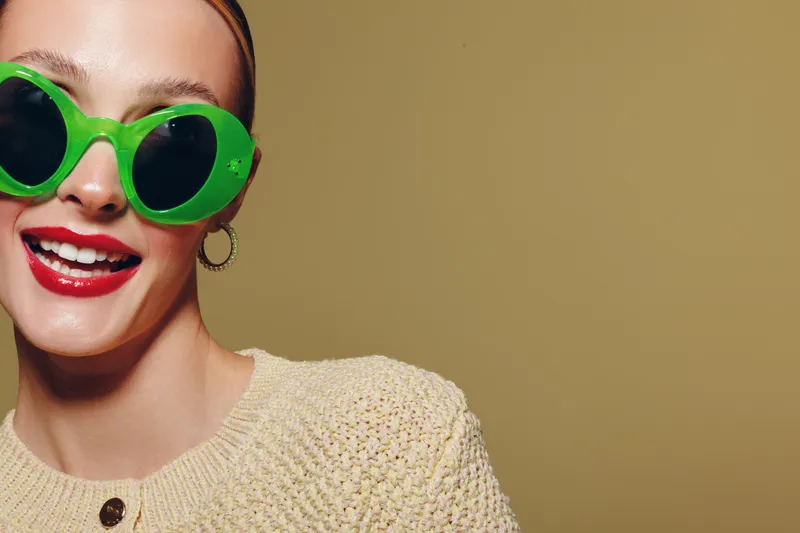 Smiling woman wearing large green sunglasses and red lipstick poses against beige background. She wears a knitted cream sweater and gold hoop earrings. Concept of fashion, style, and summer fun.