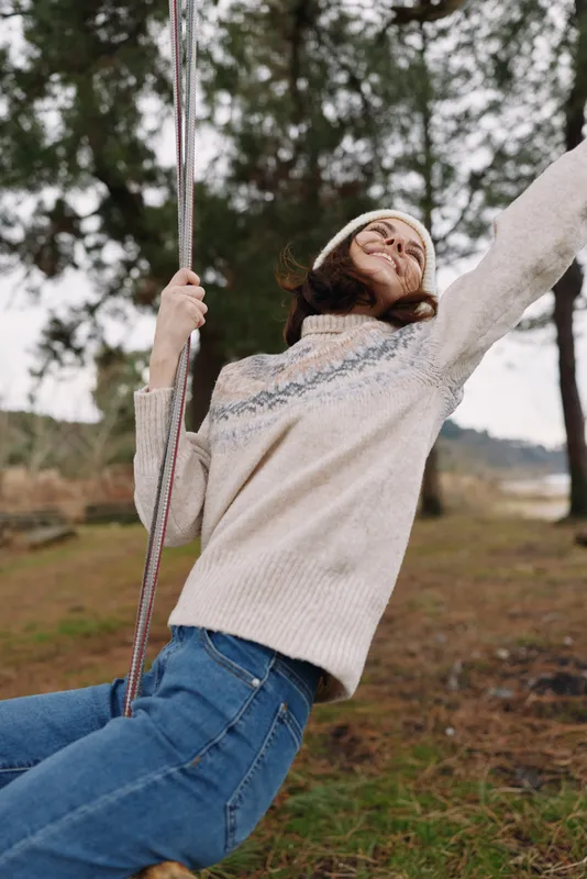 Woman on a swing outdoors in a cozy sweater, enjoying a sunny moment in nature with a wide smile and open arms, feeling carefree.
