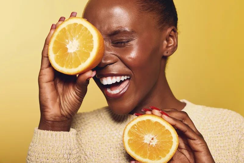 Happy African woman with dark skin holds two orange halves near face smiling with eyes closed on yellow background. Concept of healthy lifestyle, fruit, joy, and natural beauty.