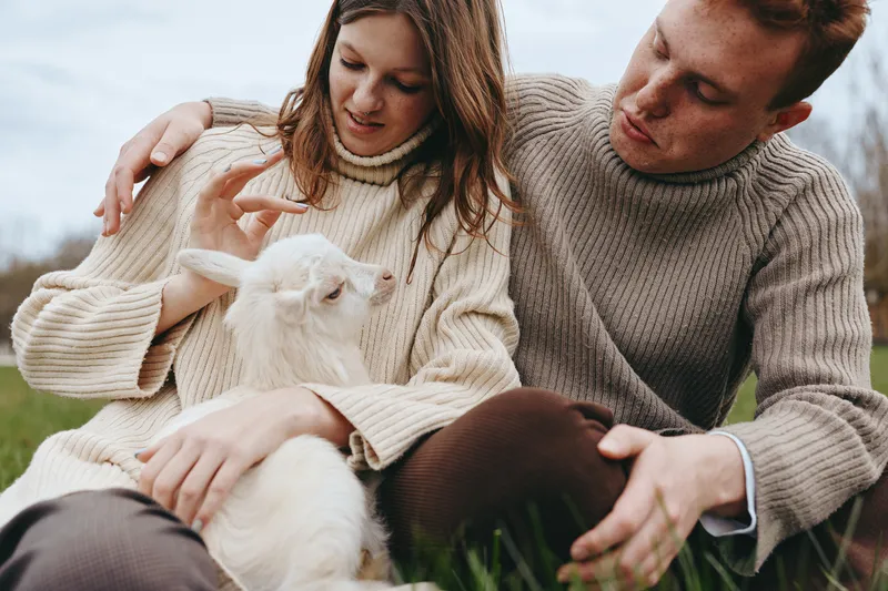 A couple shares a warm moment outdoors with a gentle goat kid, enjoying countryside life in sweaters, expressing affection and a relaxed mood among grass and meadow, capturing a candid family scene