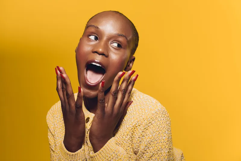 Excited young black woman with dark skin wearing yellow sweater expressing surprise with open mouth and hands near face. Studio portrait on bright yellow background. Concept for emotion and joy.