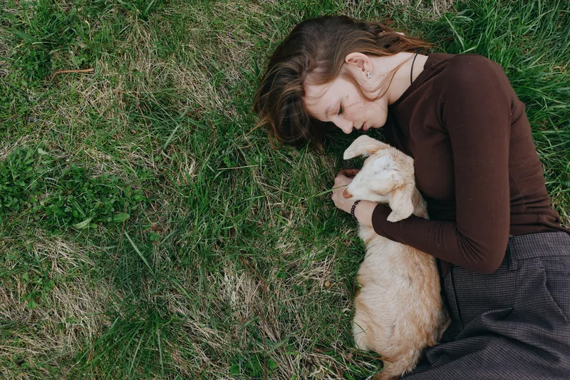 Young woman lying on grass gently hugging a baby goat in a cozy brown sweater, enjoying a peaceful moment outdoors in a natural lifestyle setting.