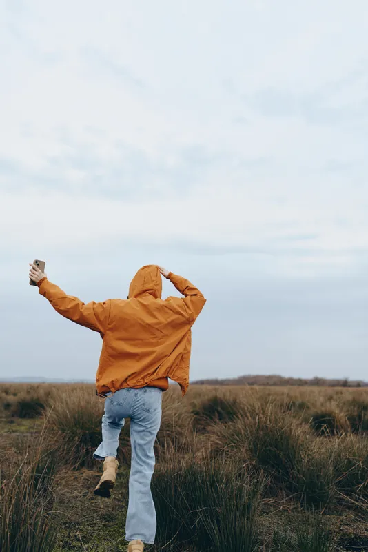Woman outdoor field sky lifestyle jacket clouds nature horizon, woman from behind walks in grassy countryside, orange hooded jacket, hands up, cloudy sky, calm lifestyle moment today