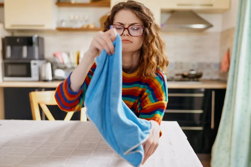 Woman ironing laundry fabric at home in a bright kitchen, wearing a colorful striped sweater and glasses. Close-up on a calm, focused moment of domestic chores, highlighting home life, routine, and