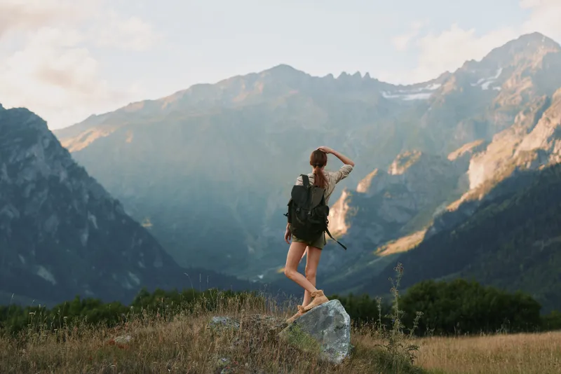 Young woman with backpack stands on a rock in a mountain valley, looking toward distant peaks in a sunlit alpine landscape, feeling freedom and adventure as a gentle breeze rustles grass